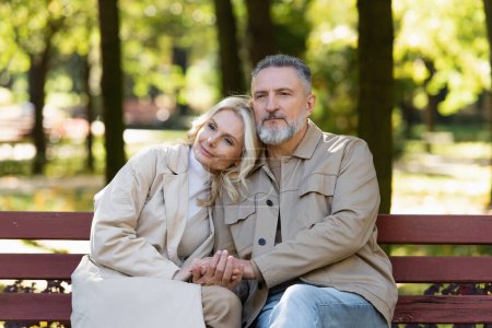 Middle aged couple holding hands while sitting on bench in park 