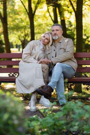 Blonde woman holding hand of mature husband while sitting on bench in spring park 