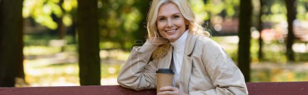 Positive Frau blickt in die Kamera und hält Coffee to go im Frühlingspark, Banner 