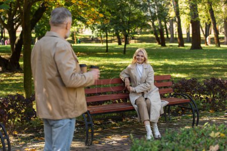 Téléchargez les photos : Femme blonde souriante regardant mari flou avec du café pour aller dans le parc de printemps - en image libre de droit
