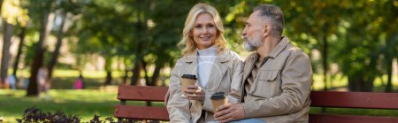 Photo for Smiling blonde woman holding takeaway coffee near husband on bench in spring park, banner - Royalty Free Image