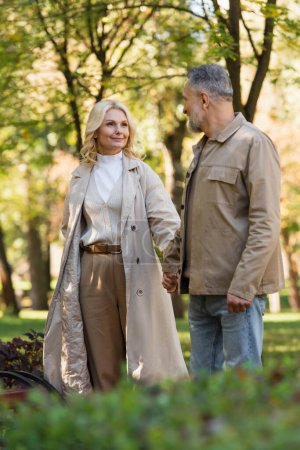 Positive blonde Frau im Trenchcoat hält Hand ihres Mannes im Park 