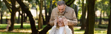 Mature man covering eyes of cheerful wife in park, banner 