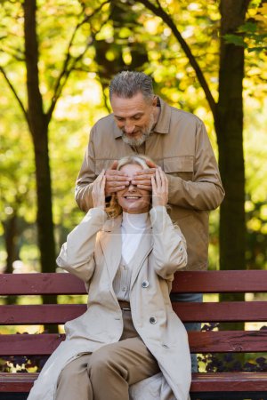 Positive mature man covering eyes of wife sitting on bench in park 