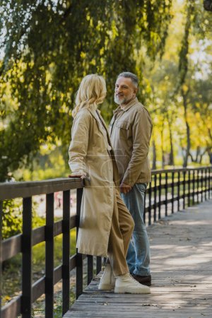Middle aged man smiling at blonde wife in trench coat while standing on bridge in park 