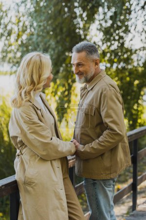 Lächelnder älterer Mann hält die Hand seiner blonden Frau, während er auf einer Brücke im Park steht 