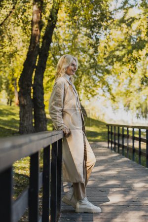 Mujer rubia madura en gabardina sonriendo mientras está de pie en el puente en el parque 
