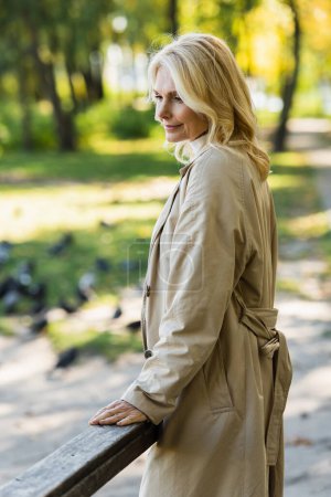 Überglückliche blonde Frau im Trenchcoat steht auf Brücke im Frühlingspark 