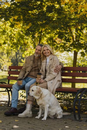 Foto de Sonriente hombre abrazando esposa con café para ir y sentado cerca de labrador en parque - Imagen libre de derechos