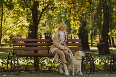 Smiling mature woman holding coffee to go while sitting on bench near labrador in park 