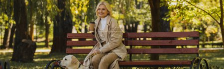 Cheerful blonde woman holding coffee to go while spending time with labrador in park, banner 