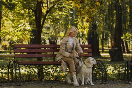 Positive woman in trench coat holding coffee to go while sitting on bench near labrador in park 