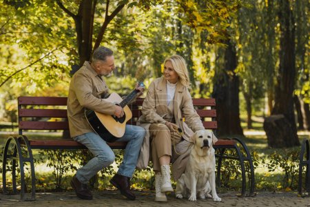 Hombre maduro tocando la guitarra acústica cerca de sonriente esposa y labrador en el parque 