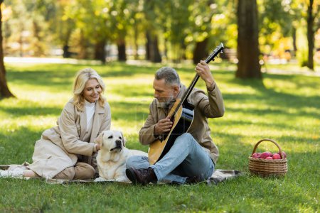 Photo for Bearded middle aged man playing acoustic guitar near happy wife and labrador dog during picnic in park - Royalty Free Image