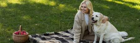 fröhliche Frau mittleren Alters mit blonden Haaren, die Labrador-Hund kuschelt, während sie auf Decke im Park sitzt, Banner 