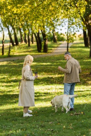 Blonde Frau mittleren Alters hält Kaffee, um mit glücklichem Mann und Labrador im Park spazieren zu gehen 