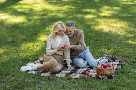 happy husband and wife holding red apple and sitting on blanket during picnic in park 