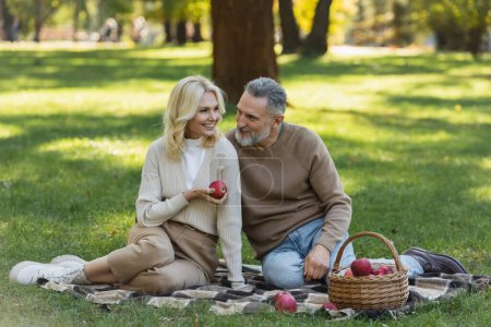 cheerful middle aged woman holding ripe apple near bearded husband during picnic in park 