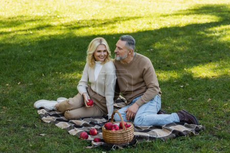 pleased middle aged woman holding ripe apple near bearded husband during picnic in park 