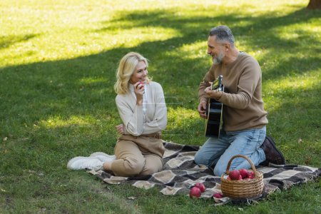 heureux homme d'âge moyen avec barbe grise jouer de la guitare acoustique près de femme insouciante avec pomme pendant le pique-nique dans le parc 