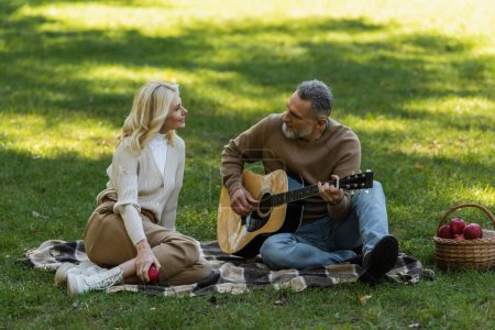 middle aged man with grey beard playing acoustic guitar near charming wife during picnic in park 