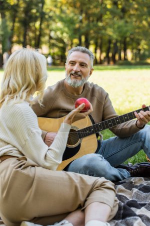 joyful middle aged man with grey beard playing acoustic guitar near blonde wife with red apple during picnic in park 