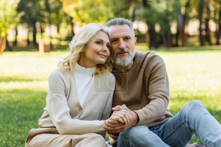 Téléchargez les photos : Portrait de charmant couple d'âge moyen tenant la main tout en étant assis ensemble dans un parc verdoyant au printemps - en image libre de droit