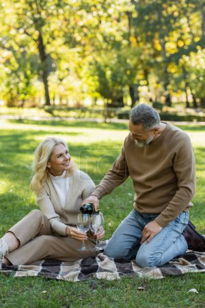 hombre de mediana edad verter vino en el vaso cerca de esposa alegre durante el picnic en el parque 