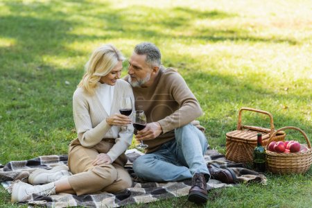 Photo for Bearded middle aged man clinking glasses of wine with blonde and happy wife during picnic - Royalty Free Image