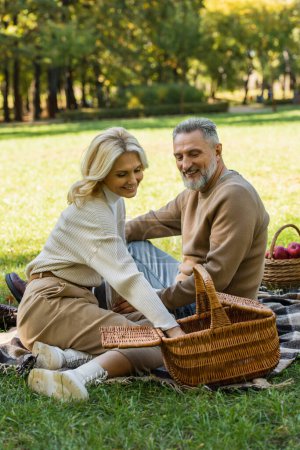 happy middle aged woman reaching food in picnic basket near cheerful husband in park 