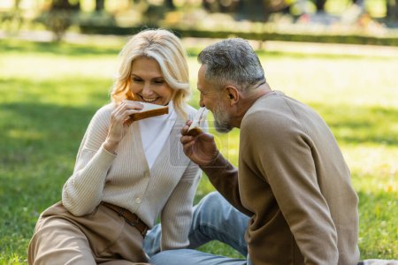 feliz pareja de mediana edad comiendo sabrosos sándwiches de club durante el picnic en el parque verde 