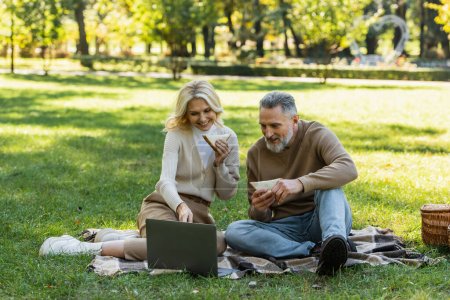 happy middle aged couple eating tasty club sandwiches and watching movie on laptop during picnic in green park 