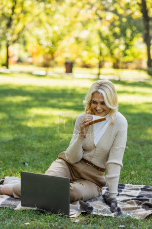 glückliche Frau mittleren Alters mit blonden Haaren, die Sandwich isst und während eines Picknicks im Park einen Film auf dem Laptop anschaut 