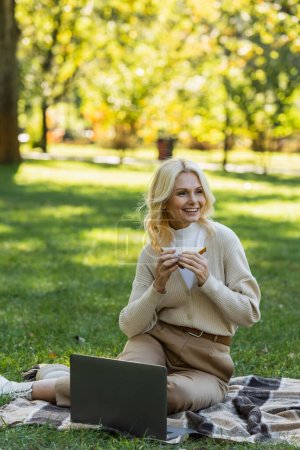 mujer de mediana edad feliz comer sándwich club y sentado en la manta cerca de la computadora portátil durante el picnic en el parque 