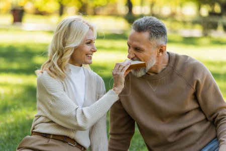 happy middle aged woman feeding bearded husband with tasty sandwich 