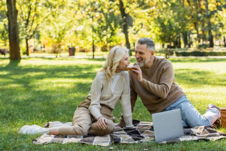 happy middle aged man feeding blonde wife with tasty sandwich during picnic in park 