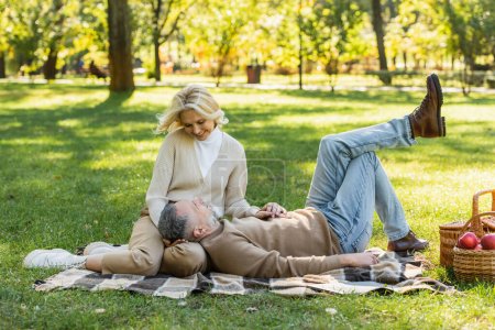 bearded middle aged man lying on laps of happy wife during picnic in park 