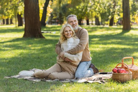 happy middle aged man with grey beard hugging charming blonde wife during picnic in park 