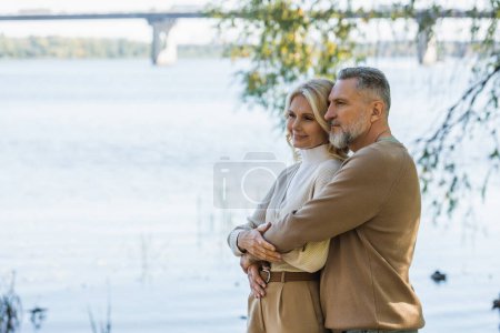 Photo for Cheerful middle aged man with grey beard hugging happy blonde wife near river in park - Royalty Free Image