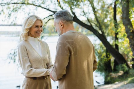 Photo for Happy middle aged woman holding hands with husband while looking at each other near lake in park - Royalty Free Image