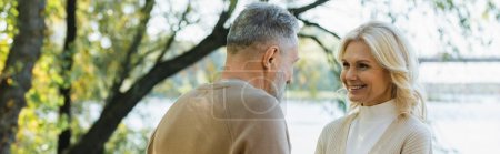 Photo for Happy middle aged woman looking at bearded husband near lake in park, banner - Royalty Free Image