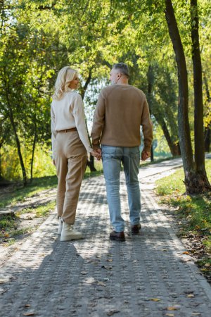 volle Länge des stilvollen Paares mittleren Alters Händchen haltend beim gemeinsamen Spaziergang im grünen Park 