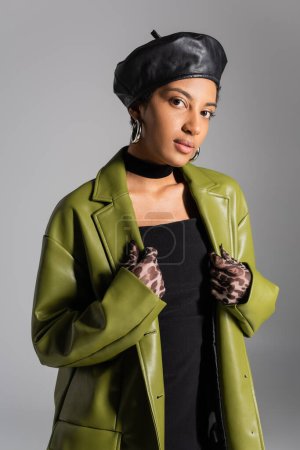 Stilvolles afrikanisch-amerikanisches Model in Baskenmütze und Lederjacke steht isoliert auf grau 