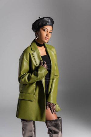 Fashionable african american model in leather beret and coat posing isolated on grey 