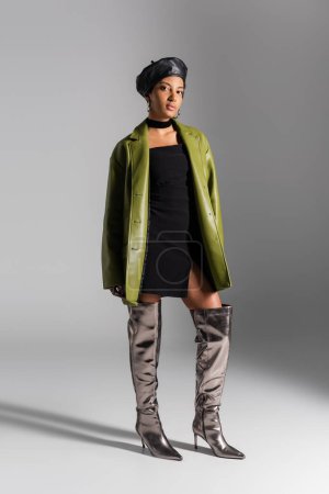 Volle Länge des stylischen afrikanisch-amerikanischen Modells in Stiefeln und Ledermantel auf grauem Hintergrund