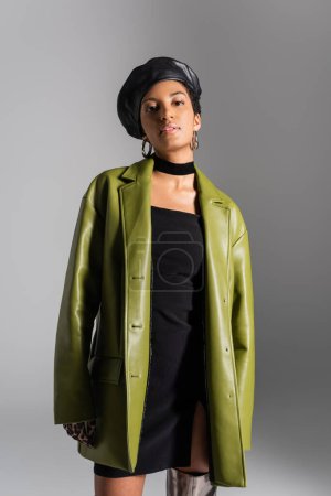 Junge afrikanisch-amerikanische Frau in Lederbarett und Mantel blickt vereinzelt in die Kamera auf grau 
