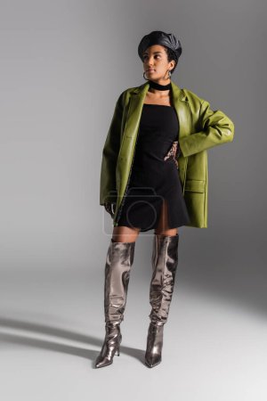 Fashionable african american woman in shiny boots and leather coat posing on grey background