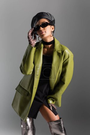Modèle afro-américain à la mode en manteau de cuir touchant les lunettes de soleil isolées sur gris 