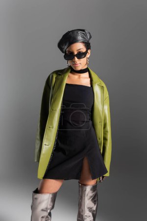 Trendy african american model in leather coat and shiny boots isolated on grey 