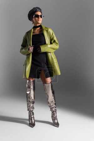 Longitud completa del modelo afroamericano de moda en abrigo de cuero y gafas de sol sobre fondo gris
